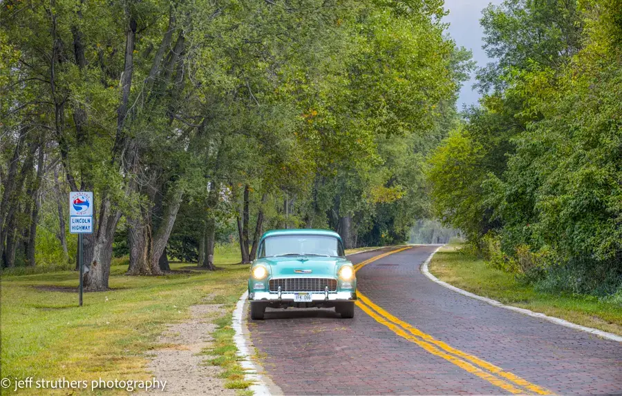 A vintage car drives down the road in the woods.